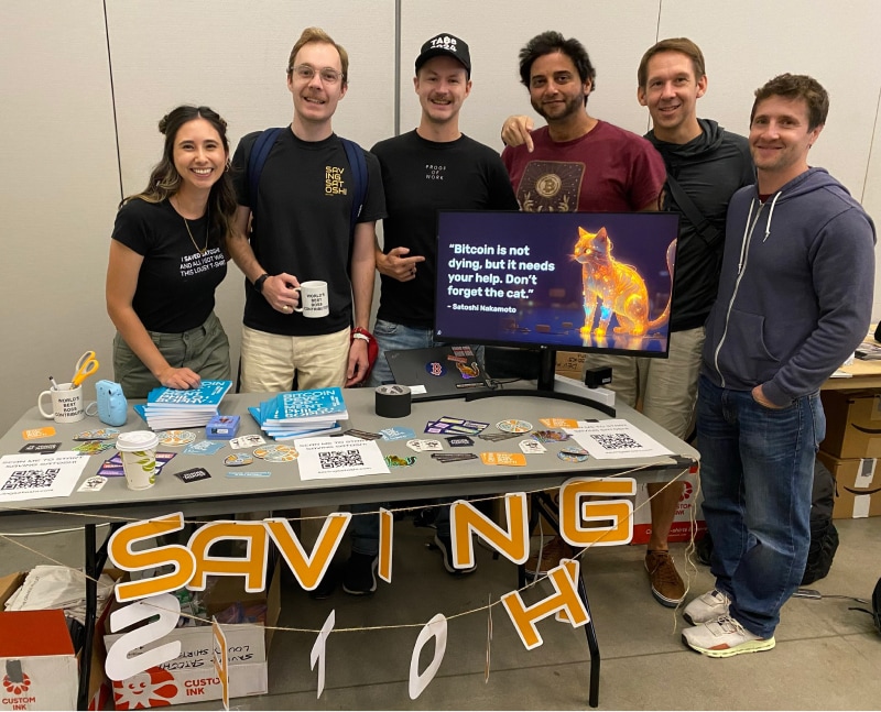 The Saving Satoshi team standing behind their conference booth, which displays merchandise, stickers, and a monitor showing the glowing cat logo. A 'SAVING SATOSHI' banner decorates the table front.